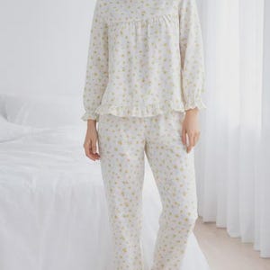 May include: A two-piece pajama set in a white fabric with a delicate yellow floral pattern. The long-sleeved top has a round neckline and ruffle trim, paired with matching pants. The pajamas are designed for comfort and relaxation.