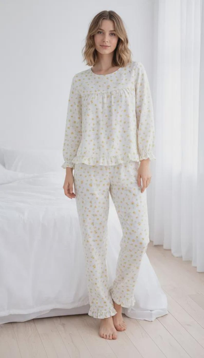 May include: A two-piece pajama set in a white fabric with a delicate yellow floral pattern. The long-sleeved top has a round neckline and ruffle trim, paired with matching pants. The pajamas are designed for comfort and relaxation.