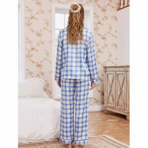Op de afbeelding: Een blauw-witte geruite pyjamaset. De top met lange mouwen en de broek hebben een klassiek ruitpatroon. De pyjama's hebben rode biezen op de manchetten en zomen. De persoon draagt de pyjama in een slaapkamer.