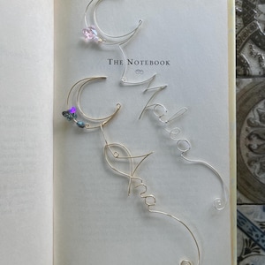 May include: Two wire bookmarks resting on an open book titled The Notebook. One bookmark is silver with a crescent moon and a pink bead, the other is gold with a crescent moon and dark beads. Both feature cursive wire lettering.