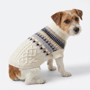 May include: A cream-colored dog sweater with a cable knit design on the front and a patterned yoke in navy, tan, and gray. The sweater has a turtleneck and is designed for a small dog breed.
