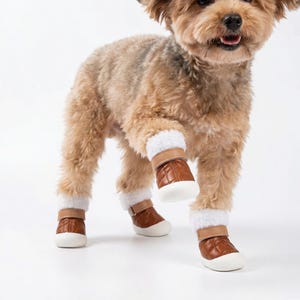 Op de afbeelding: Een kleine hond met vier bruine leren hondenlaarzen met witte imitatiebontafwerking en witte zolen. De laarzen hebben een gewatteerd patroon en verstelbare bandjes. De hond staat op een witte ondergrond.