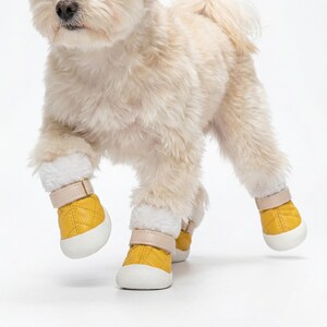 Op de afbeelding: Een kleine hond draagt vier gele hondenlaarzen met witte zolen en beige bandjes. De laarzen hebben een gewatteerd patroon en een zachte, witte voering. De hond loopt op een witte ondergrond.