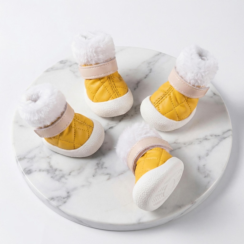 Op de afbeelding: Vier gele gewatteerde hondenlaarzen met witte zolen en een pluizige witte rand. Elke laars heeft een beige band. De laarzen zijn gerangschikt op een wit marmerachtig oppervlak. Deze laarzen zijn ontworpen om de poten van een hond te beschermen.