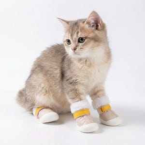 Op de afbeelding: Een lichtbruin katje draagt beige en gele dierenlaarsjes met witte imitatiebontafwerking. De laarzen hebben een gewatteerd ontwerp en een gele band. De kat zit op een witte achtergrond.