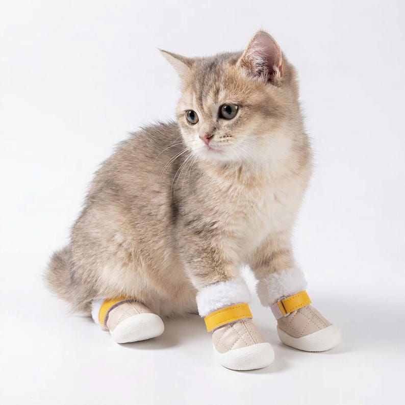 Op de afbeelding: Een lichtbruin katje draagt beige en gele dierenlaarsjes met witte imitatiebontafwerking. De laarzen hebben een gewatteerd ontwerp en een gele band. De kat zit op een witte achtergrond.