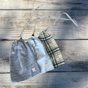 Peut inclure: Trois sacs à cordon de serrage aux motifs variés : rayures bleues et blanches, bleu clair uni et carreaux bleus et verts. Chaque sac est doté d'un cordon de serrage rayé assorti et d'une petite étiquette. Les sacs sont disposés sur une surface en bois vieilli.