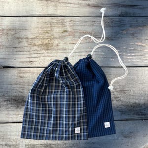 May include: Two drawstring bags with a plaid pattern in shades of blue, black, and white. The bags have white drawstrings and small white labels. The bags are made of fabric and are likely reusable.