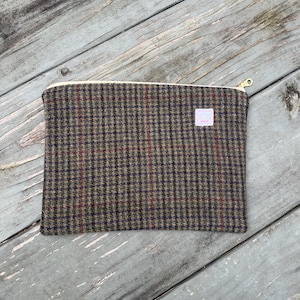 May include: A rectangular, zippered pouch made of a brown, gray, and red plaid fabric. The pouch has a cream-colored zipper and a small, light pink tag on the front. The background is a weathered, gray wooden surface.