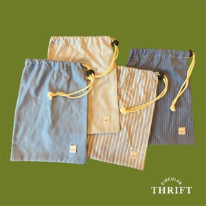 May include: Four rectangular drawstring bags in shades of light blue, cream, and blue-and-white stripes. Each bag features a cream-coloured rope drawstring with a black toggle and a small white rectangular label with the text CIRCULAR THRIFT.