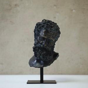May include: Bronze bust of a bearded man with curly hair, mounted on a black metal stand. The sculpture has a weathered, antique appearance, with detailed facial features and a prominent beard. The bust is set against a neutral background.
