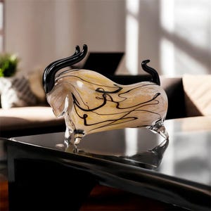 Könnte beinhalten: Dekorative Glas-Stierskulptur mit cremefarbenem und gelbem Körper, schwarzen Hörnern und schwarzen abstrakten Linien. Der Stier wird auf einer schwarzen Oberfläche ausgestellt, die seine transparenten Glasbeine und seinen Körper zeigt.