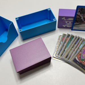 May include: An assortment of card storage boxes and card sleeves in various colors. Includes blue and purple boxes, a stack of purple card sleeves, and a deck of cards. A single card is displayed in a blue frame.