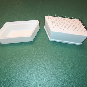 May include: Two white, diamond-shaped containers are on a green surface. One is empty, the other holds many thin, silver needles. The containers are likely plastic and used for a craft or medical purpose. The needles are likely used for acupuncture.
