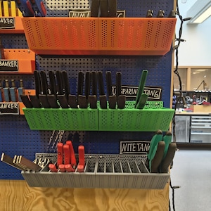 May include: A workshop wall display featuring various tools organized in colorful, perforated plastic bins. The bins are orange, green, and gray, and hold tools like hammers, pliers, and measuring tools. Labels identify the tools.