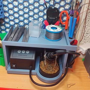 May include: A blue and gray electronics workstation with various tools and components. Includes a soldering iron, solder, tweezers, and an ESD SAFE device. The background features a blue honeycomb-patterned wall.