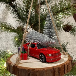 May include: A red toy car ornament with a silver tree on a wooden slice, decorated with small gift boxes. The ornament hangs from a Christmas tree with pine needles and pine cones.