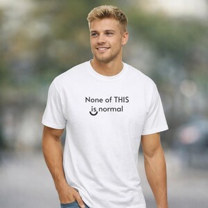 May include: White t-shirt with the text "None of THIS is normal" in black. The shirt has a crew neck and short sleeves. The person is wearing blue jeans.