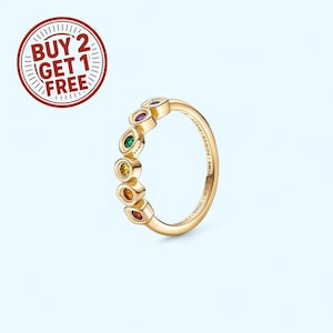 Puede incluir: Un anillo dorado con una serie de gemas de colores en un patrón de arcoíris. El anillo tiene un acabado liso y pulido y está sobre un fondo azul claro. La banda del anillo tiene las palabras "TIME - MIND - SOUL - SPACE - POWER" grabadas.