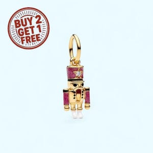 May include: A gold-colored charm in the shape of a toy soldier, with a pink glittery hat and accents. The charm has a star-shaped gem and white boots. The charm is designed to be a pendant or a decorative element for jewelry.