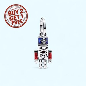 May include: A silver charm in the shape of a toy soldier, featuring a blue and red design with a star on the hat. The charm has a loop for attaching to a bracelet or necklace. The image also includes a red and white circular graphic with the text "BUY 2 GET 1 FREE".