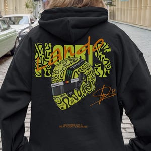 May include: Black hoodie featuring a neon green and black design of a racing helmet and the name "NORRIS" in a stylized font. The hoodie also includes a signature and the text "BEAT YOUR TEAM MATE".