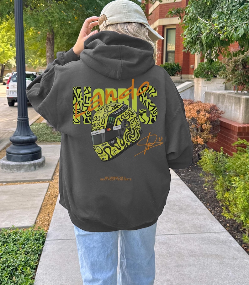 May include: Dark gray hoodie with a graphic design on the back. The design features the word "Norris" in orange and a neon green and black helmet illustration. The hoodie also has a signature and the text "BELIEVERS ONLY. MEET YOUR TEAM MATE."