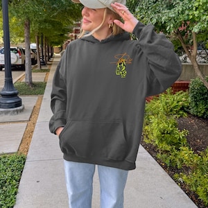 May include: Dark gray hoodie with a kangaroo pocket and a drawstring hood. The hoodie features a small, stylized design in yellow and orange on the upper left chest. The person in the image is wearing a beige baseball cap and light blue jeans.