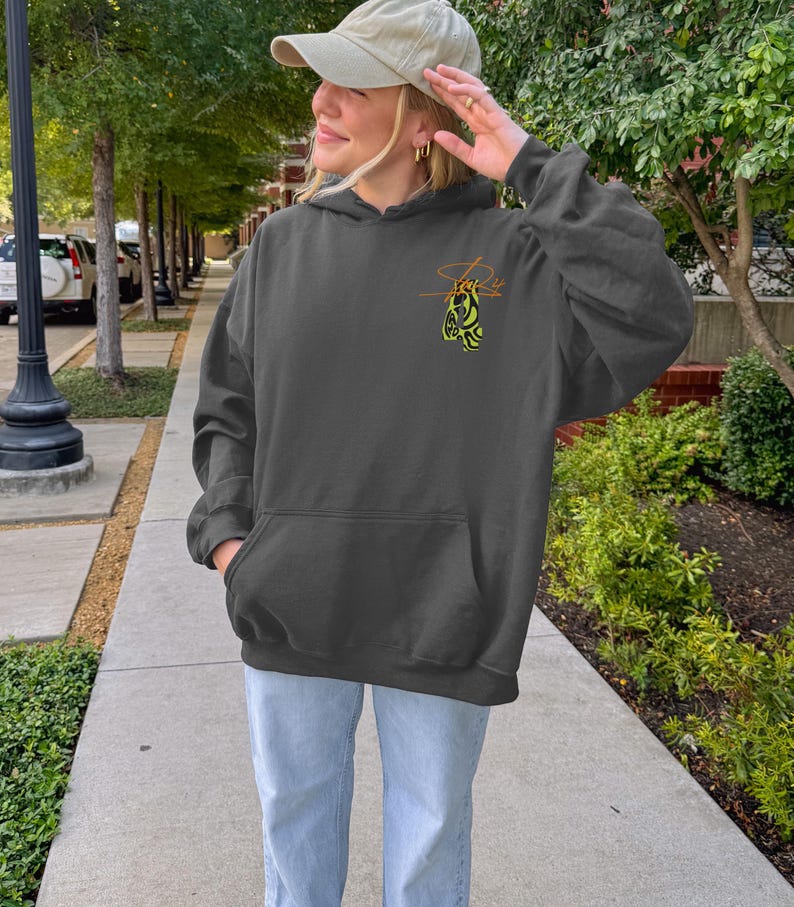 May include: Dark gray hoodie with a kangaroo pocket and a drawstring hood. The hoodie features a small, stylized design in yellow and orange on the upper left chest. The person in the image is wearing a beige baseball cap and light blue jeans.