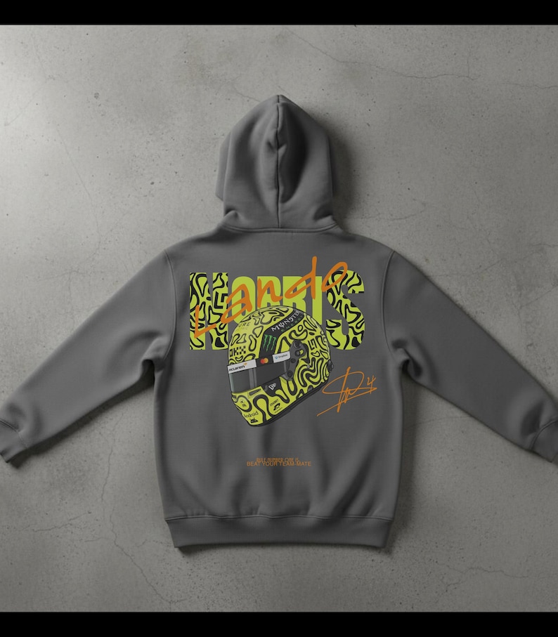May include: A gray hoodie featuring a graphic of a racing helmet with the name "Norris" in yellow and black. The hoodie has a hood and long sleeves. The back of the hoodie displays the helmet design and signature.