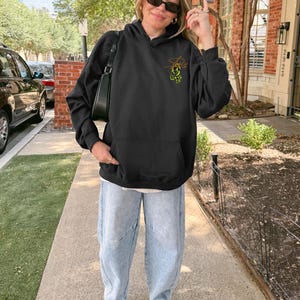 May include: Black hooded sweatshirt with a front pocket and a small graphic on the upper left chest. The graphic is a stylized design with orange and green accents. The person is wearing light blue jeans and white sneakers.