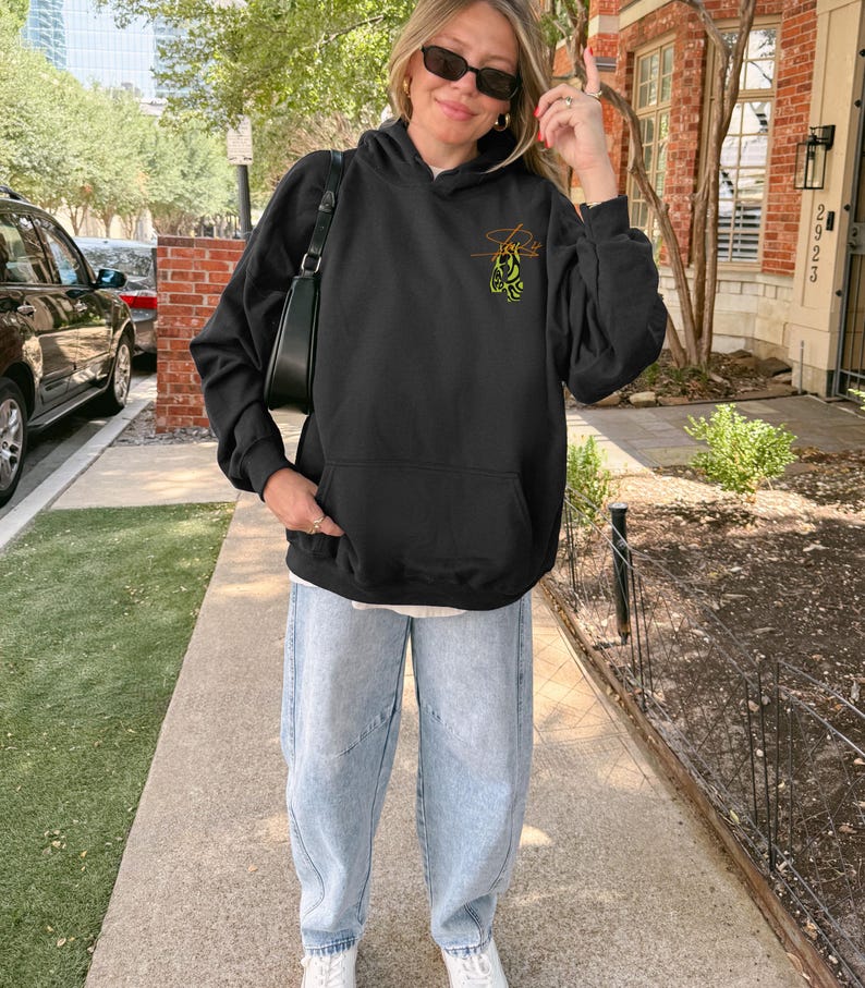 May include: Black hooded sweatshirt with a front pocket and a small graphic on the upper left chest. The graphic is a stylized design with orange and green accents. The person is wearing light blue jeans and white sneakers.