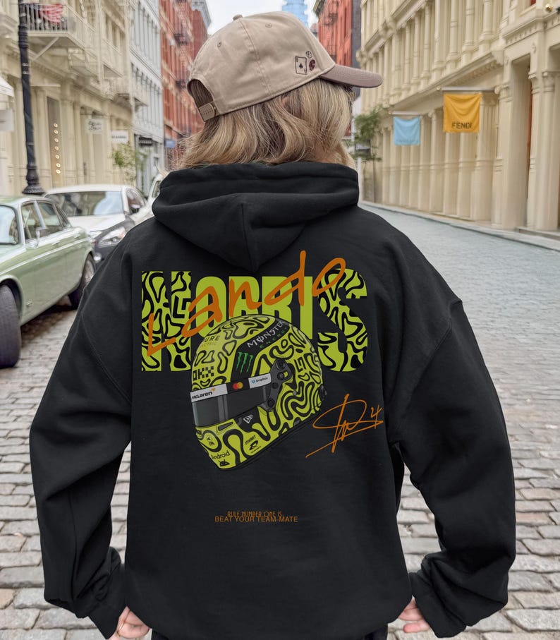 May include: Black hoodie featuring a neon green and black design of a racing helmet and the name "NORRIS" in a stylized font. The hoodie also includes a signature and the text "BEAT YOUR TEAM MATE".
