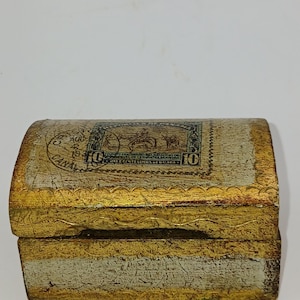 May include: A small, vintage wooden chest with a curved lid. The chest is painted in a distressed style with gold and off-white tones. A faded stamp design is visible on the lid, adding to its antique aesthetic.