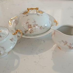 May include: A white porcelain serving set with a floral pattern and gold accents. The set includes a covered dish, a creamer, and a sugar bowl. The floral pattern features pink and green flowers.