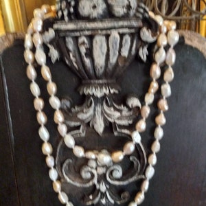 May include: A double strand of cream and white pearl beads draped over a decorative, ornate, black and white, carved wooden object.