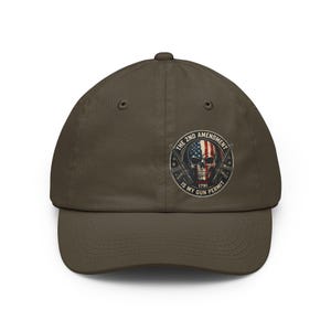 Peut inclure: Casquette de baseball vert olive avec un motif de crâne intégrant le drapeau américain et des fusils croisés. Le texte indique "THE 2ND AMENDMENT IS MY GUN PERMIT 1791".