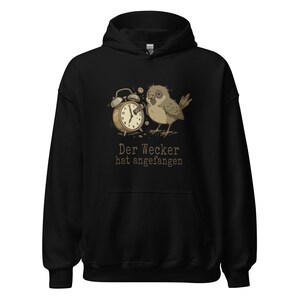Könnte beinhalten: Schwarzer Kapuzenpullover mit einem Motiv eines müden Vogels und eines Weckers. Der deutsche Text "Der Wecker hat angefangen" ist darunter gedruckt. Der Hoodie hat eine Kapuze mit Kordelzug und eine Vordertasche.