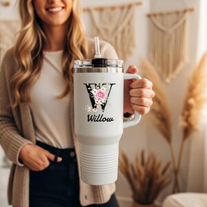 May include: A white insulated travel mug with a handle, decorated with a floral "V" and the name "Willow". The mug has a black lid and a straw. The mug is designed to keep drinks hot or cold.