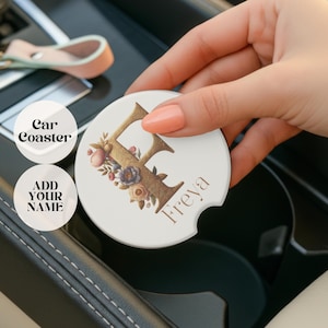 Pode incluir: Um porta-copos branco para carro com um design floral e a letra "F" dourada. O nome "Freya" é impresso em uma fonte cursiva. O porta-copos está sendo segurado perto do porta-copos de um carro. O texto na imagem diz "Car Coaster" e "Add Your Name".
