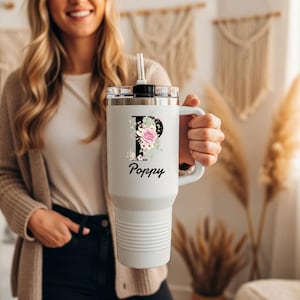 Può includere: Bicchiere termico bianco con coperchio e manico neri. Il bicchiere presenta un motivo floreale con la lettera "P" e il nome "Poppy" in corsivo. Il bicchiere ha una cannuccia ed è tenuto da una persona.
