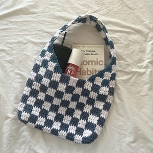May include: A handmade crochet tote bag in a blue and white checkerboard pattern. The bag contains a black tumbler, a bottle of nail polish, and a book titled "Atomic Habits". The bag has a matching handle.