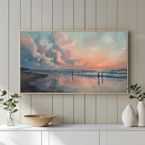 May include: A framed landscape painting depicts a beach scene at sunset. The sky is filled with pink and blue clouds, reflecting in the wet sand. Silhouettes of people stand on the shore, with gentle waves rolling in. The artwork is displayed in a light wood frame.