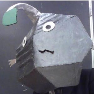 Puede incluir: Una persona con un disfraz de rape hecho a mano. El disfraz presenta una cabeza geométrica gris con acentos de ojos blancos, una boca negra y una aleta verde. La persona lleva un traje y una corbata negros.