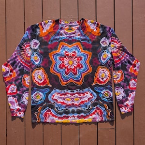 May include: A long-sleeve crew neck shirt featuring a black base with a vibrant tie-dye pattern. The design includes a central star-shaped motif surrounded by various organic, multi-colored shapes in shades of red, orange, yellow, blue, and purple.