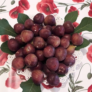 Può includere: Una ciotola bianca e traforata piena di fichi viola maturi. I fichi sono disposti su foglie verdi di fico. La ciotola è appoggiata su una tovaglia floreale con papaveri rossi.
