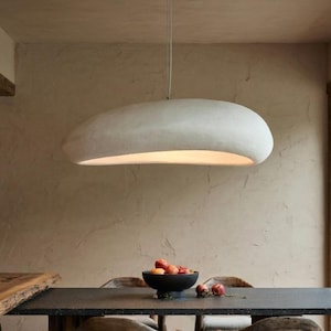 May include: A modern dining room featuring a large, dark rectangular table with wooden chairs. A large, white, oval-shaped light fixture hangs above the table. A marble stool and a view of a Japanese garden are also visible.