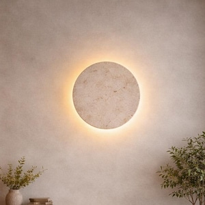 Puede incluir: Una oficina en casa moderna con un escritorio de color claro, una computadora portátil y una silla de cuero. Una lámpara de pared redonda e iluminada está centrada sobre un mueble de madera. Un pequeño árbol en maceta y una estantería completan el espacio.