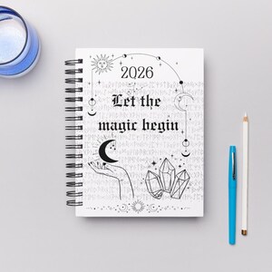 May include: A spiral-bound 2026 planner with the text "Let the magic begin" in a gothic font. The cover features a sun, moon, crystals, and hand-drawn illustrations in black and white. A blue pen and a white pencil are next to the planner.