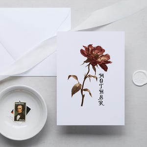 May include: A white card featuring a brown rose illustration and the word "MOTHER" in black. An envelope, a small white bowl with a stamp, a white ribbon, and a wax seal are also present.